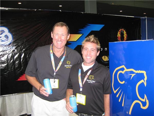 Two smiling men in black Roaring Lion polo shirts and lanyards hold blue cans in front of a blue banner with a yellow lion logo.