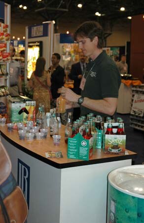 A man in a dark green polo stands behind a counter holding a bottle, with Steaz bottles, sample cups and four-pack cartons in front of him.