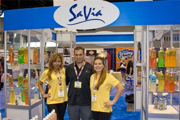A man in a black shirt stands between two women in yellow tops under a blue SaVia sign, with shelves of bottled drinks on both sides.