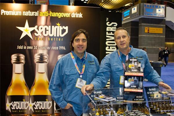 Two men in blue denim shirts stand at a display counter in front of a Security anti-hangover drink banner showing two amber bottles.