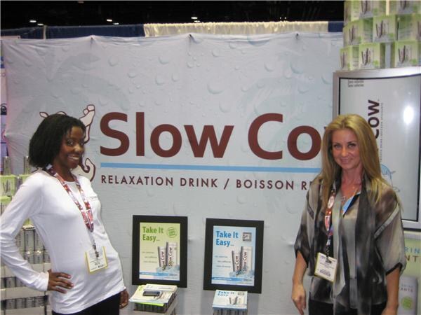 Two women stand in front of a large Slow Cow relaxation drink banner, with small Take It Easy posters and product cans on display.