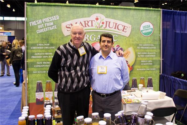 An older man in a patterned sweater and a man in a blue shirt stand before a green Smart Juice Organic banner, with juice bottles on the table.