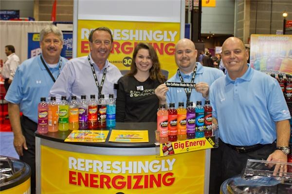 Four men and a woman stand behind a counter lined with colorful bottles under a yellow Refreshingly Energizing sign. One man holds a small sign, and a Big Energy placard is visible.