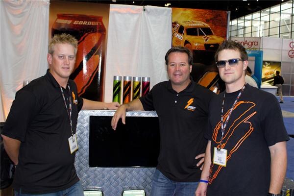 Three men in black shirts stand around a diamond-plate display with a Speed logo; behind them are a Gordon's can graphic and a race car image.