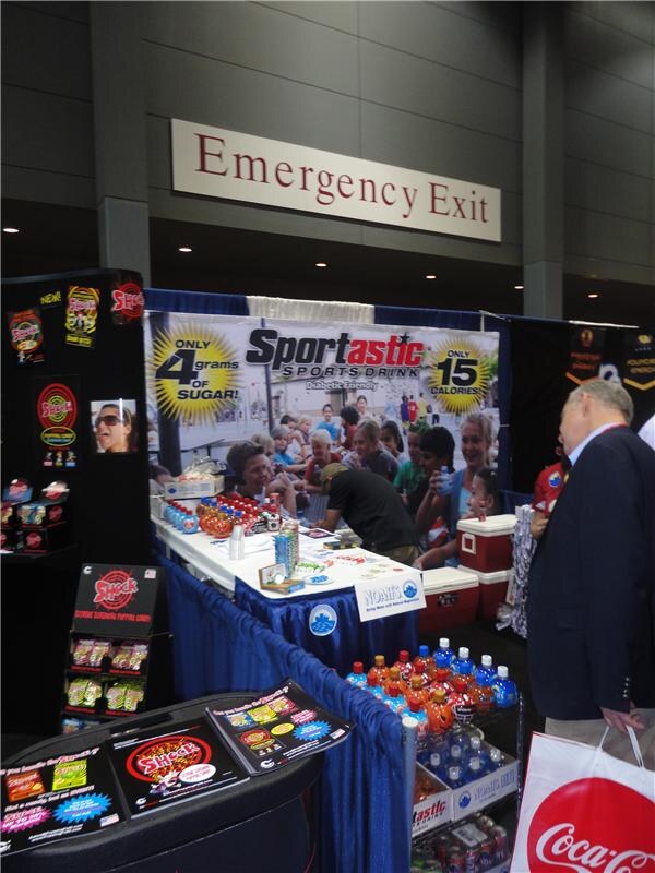 A Sportastic sports drink booth with banners and bottles. Shock and Nomi's displays are nearby, and a man holding a Coca-Cola bag looks on at right.