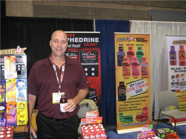 A smiling man with a lanyard holds a small bottle in a booth with Stacker 2 and 6 Hour Power banners and product displays, including a banner advertising a new extra strength grape flavor.