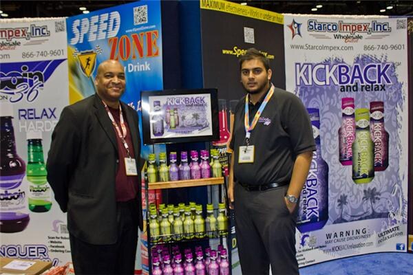 Two men stand in front of Starco Impex, Speed Zone and Kickback banners, beside a shelf of purple and green bottles.
