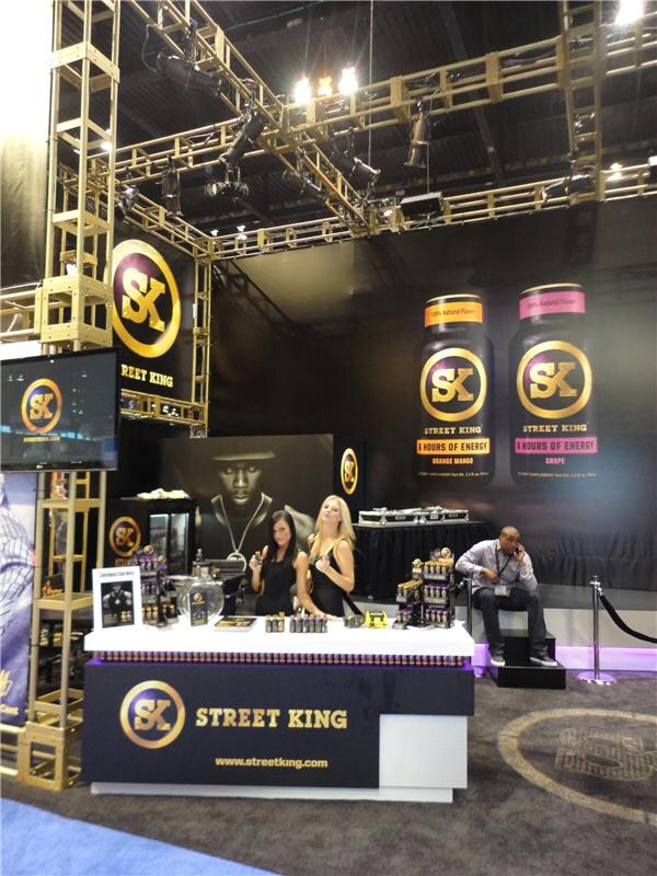 Street King booth with gold truss, large SK bottle graphics in orange mango and grape, and a counter with product displays. Two women stand behind it and a man sits on a step talking on a phone.