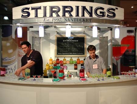 Two men stand behind a white bar under a Stirrings Est. 1997 Nantucket sign, with cocktail mixer bottles and martini glasses on the counter.