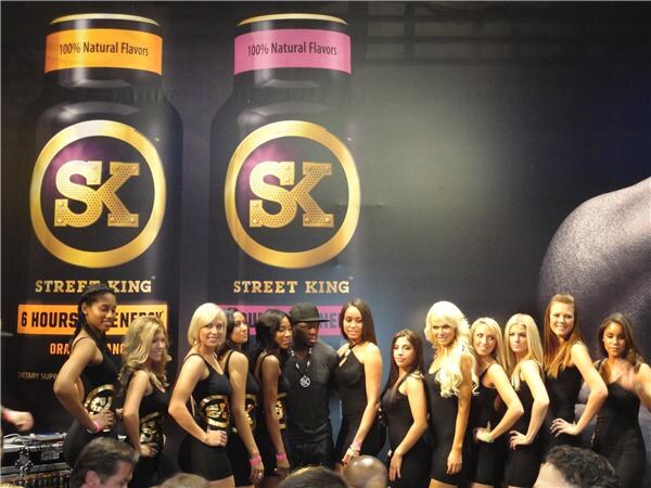 A man in a black cap and dark clothing stands among a line of women in black dresses in front of large Street King SK bottle graphics reading 100% Natural Flavors and 6 Hours of Energy.
