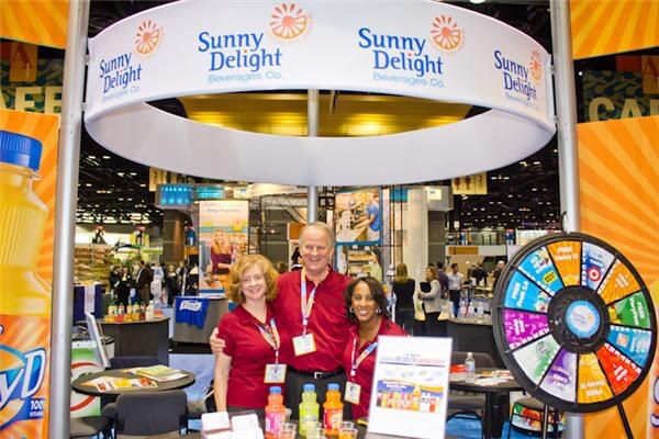 Three smiling people in red shirts and lanyards stand together under a round Sunny Delight Beverages Co. sign, with juice bottles in front and a prize wheel at right.