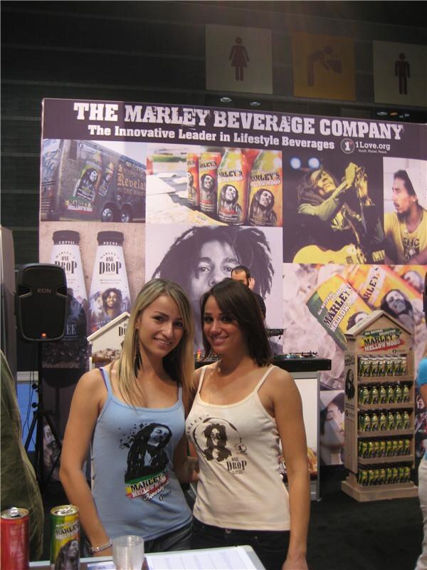 Two women in tank tops with Marley artwork pose in front of a Marley Beverage Company display showing Marley's Mellow Mood and One Drop bottles and cans, with a 1Love.org logo.
