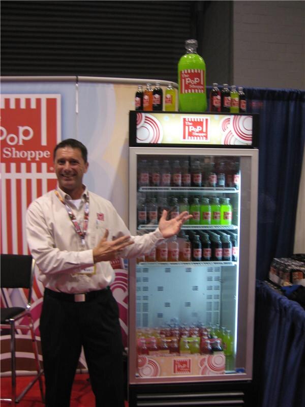 A smiling man in a light shirt and lanyard gestures toward a lit glass-door cooler of The Pop Shoppe bottled sodas, topped with a large green bottle, beside a red and white Pop Shoppe banner.