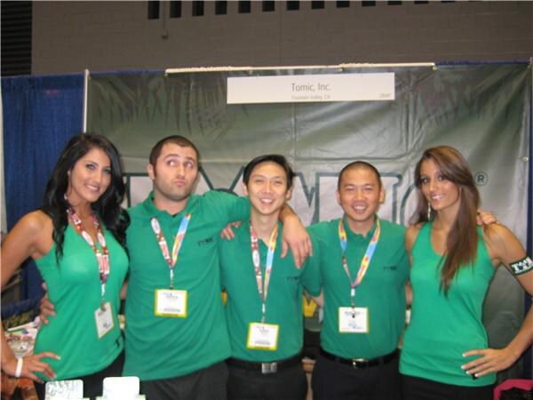 Five people in green Tomic shirts stand arm in arm in front of a banner reading Tomic, Inc., Fountain Valley, CA. Three men are in the middle, with a woman at each end, all wearing badges.