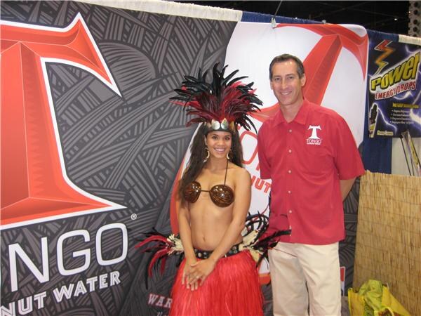 A man in a red Tongo shirt stands beside a woman in a feathered headdress, coconut-shell top and red grass skirt, in front of a Tongo coconut water banner and a Power Energy Drops sign.