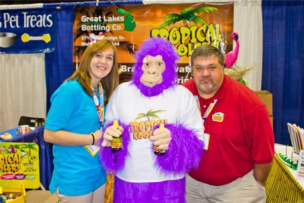 A woman in a blue shirt and a man in a red shirt flank a person in a purple gorilla costume holding two bottles, in front of a Tropical Koolers sign for Great Lakes Bottling Co.