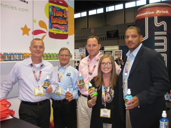 Five people in convention lanyards pose at the Tum-E Yummies booth, four holding small bottles of drink. A shelf of colorful bottles and a large Tum-E Yummies bottle graphic are behind them.