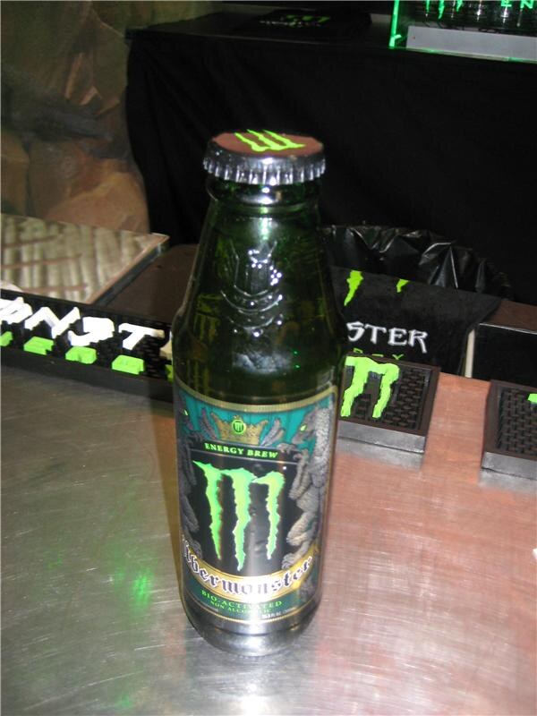 A green glass bottle of Übermonster Energy Brew from Monster Energy stands on a metal counter, with Monster-branded bar mats and a black towel with the green claw logo behind it.