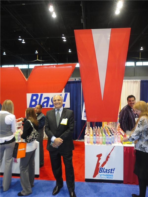 A man in a dark suit with a badge stands smiling in front of large red V sculptures and a Blast! sign. A display of colorful bottles sits on a counter, with other attendees around.