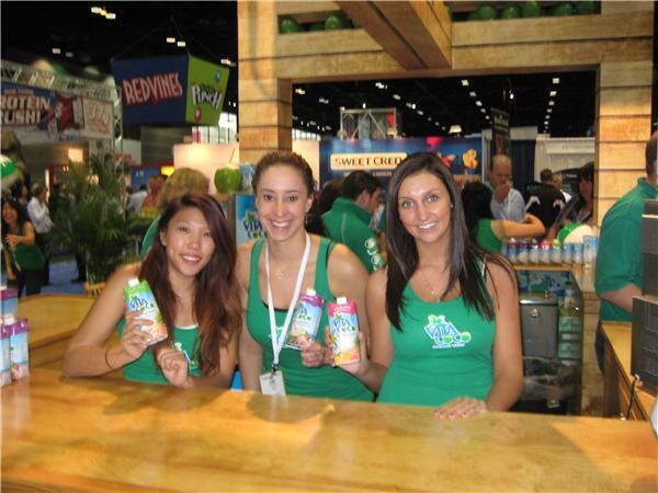 Three smiling women in green Vita Coco tank tops stand behind a wooden counter, each holding a Vita Coco coconut water pouch. Other trade show booths are visible behind them.