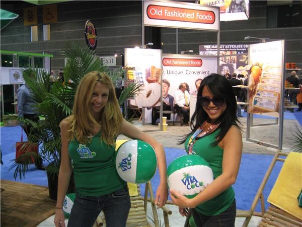 Two women in green Vita Coco tank tops hold Vita Coco beach balls on a mock beach setting with a palm plant and lounge chairs, with Old Fashioned Foods signs behind them.