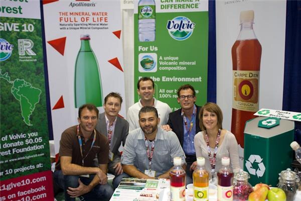 Five people pose around a booth table with Volvic, Apollinaris and Carpe Diem banners behind them. Bottles of drinks and a green recycling bin are in front.