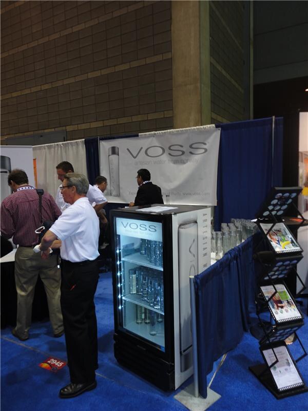 A VOSS booth with a banner reading artesan water from Norway and a lit glass-door cooler of bottles. Several men in the aisle and booth stand nearby, with brochure racks to the right.