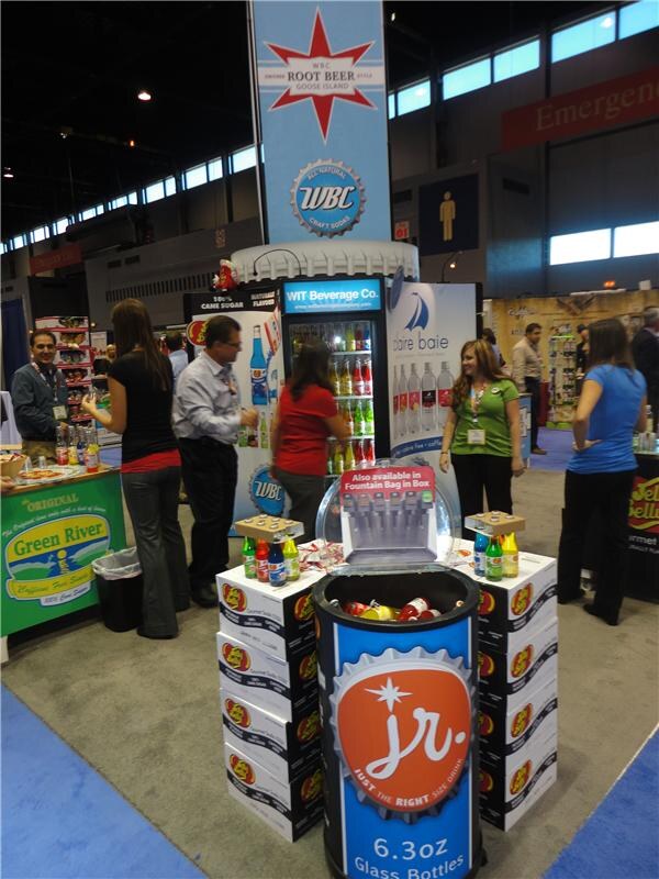 A WIT Beverage Co. booth with a WBC Root Beer Goose Island banner, a lit cooler, a Pure Baie display and a Jelly Belly Jr. bottle display. Attendees stand around, with a Green River booth at left.