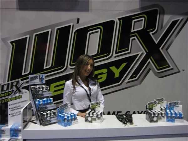 A woman in a white shirt and lanyard smiles behind a white counter displaying Worx Energy shot bottles, in front of a large black and green Worx Energy logo.