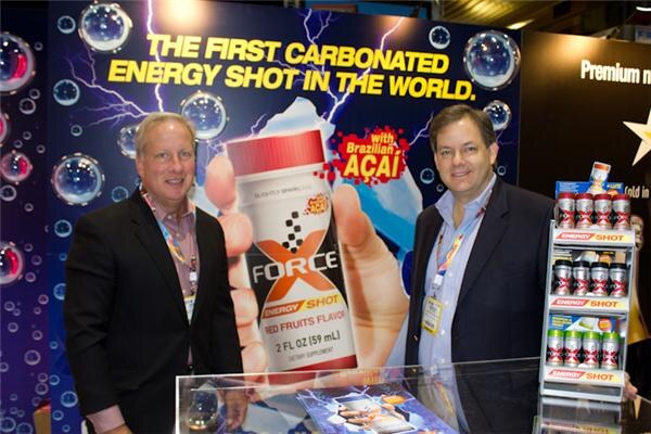 Two men in dark jackets and lanyards stand beside a large X Force Energy Shot display reading The first carbonated energy shot in the world, with a shelf of shots at right.