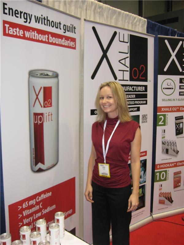 A smiling woman in a maroon top and lanyard stands between banners for Xo2 Uplift energy drink and Xhale O2. Cans of the drink sit on a table at the lower left.