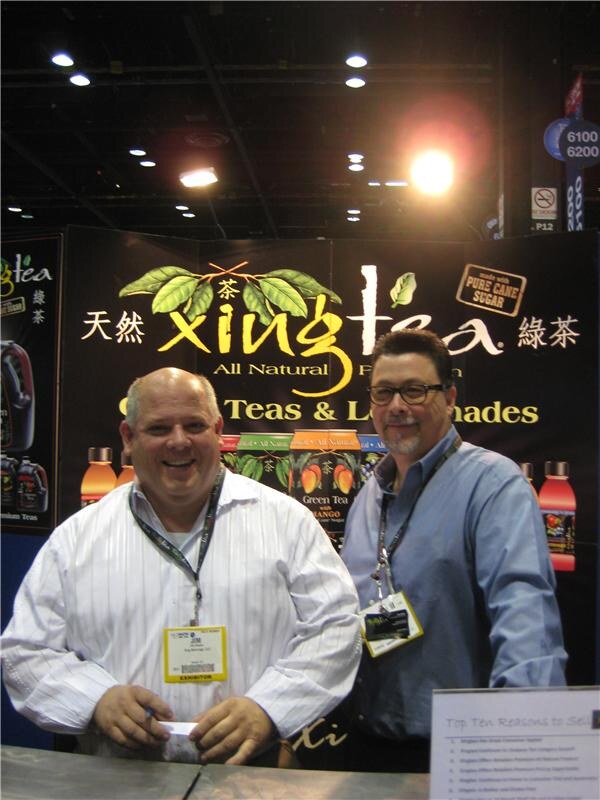 Two men with exhibitor badges and lanyards stand behind a counter in front of a black Xing Tea banner showing tea and lemonade bottles. One man smiles and holds a card.
