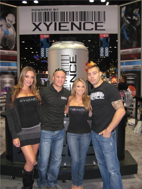 Two women in Xyience tops and two men, one in a black Xyience shirt with tattooed arm, pose in front of a Xyience booth with a large silver can display and fighter banners.