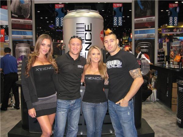 Two women and two men in Xyience clothing pose together in front of a large Xyience can display and banners. One man has a mohawk, and a tattoo is visible on his arm.