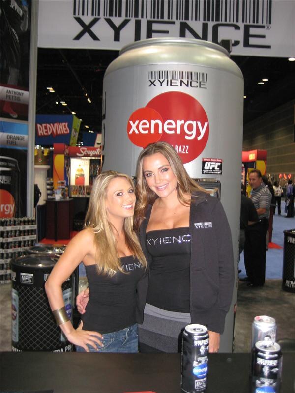 Two women in black Xyience outfits pose in front of a giant Xenergy can. Xyience cans sit on the counter, and a Redvines sign and other booths appear behind.