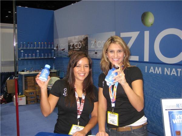 Two smiling women in black Zico shirts and lanyards hold up small blue Zico bottles in front of a blue Zico booth wall with shelves of bottles.