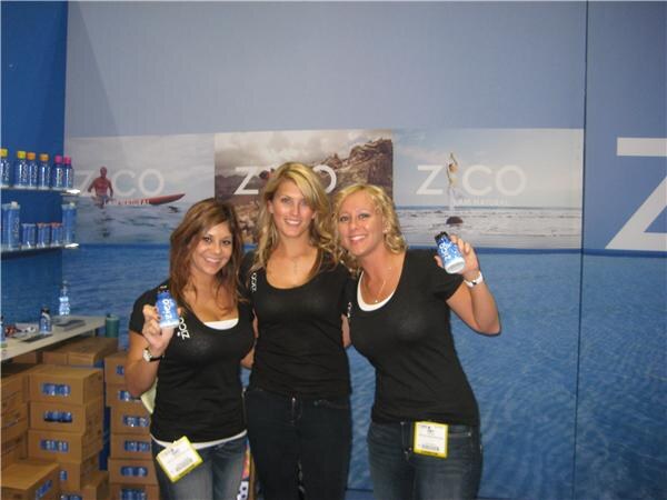Three women in black Zico shirts pose together, two holding small blue Zico bottles, in front of a blue Zico booth with shelves and stacked cartons.