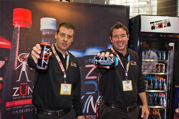 Two men in black shirts with lanyards hold out cans of Zün energy drink in front of a Zün banner and a refrigerated cooler of bottled drinks.
