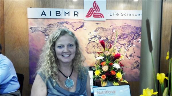A smiling woman with curly blonde hair sits in front of an AIBMR Life Sciences sign with a world map and a bouquet of flowers.