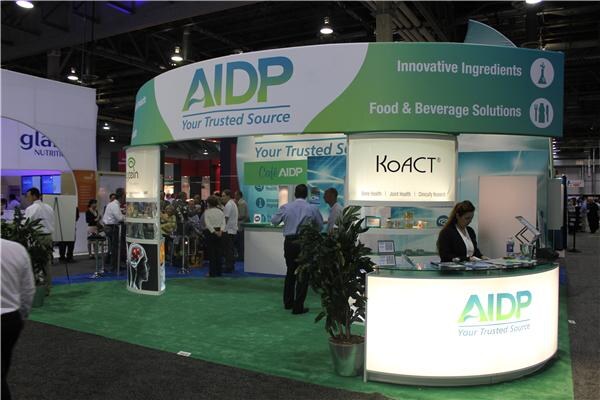 Wide view of the green and white AIDP booth with KoACT signage, a woman at a lit counter and visitors standing; a Glanbia Nutritionals booth is at left.