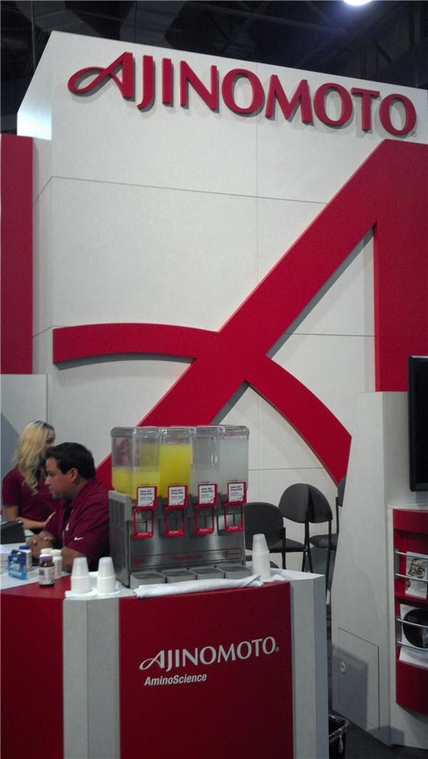 Red and white Ajinomoto AminoScience booth with a large logo, a four-tub beverage dispenser, and two staff members in red shirts seated at the counter.