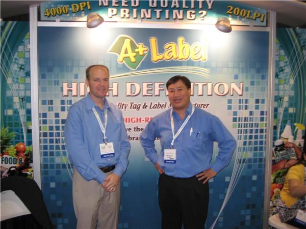 Two men in blue button-up shirts and lanyards stand in front of an A+ Label booth backdrop reading High Definition and Need Quality Printing.
