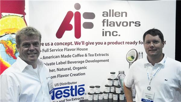 Two smiling men in white shirts stand in front of an Allen Flavors Inc. banner listing flavor services, with jars of extracts and a Nestlé US distributor logo displayed below.