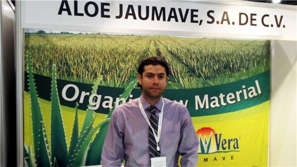 A man in a lavender shirt and striped tie wears a lanyard in front of an Aloe Jaumave, S.A. de C.V. banner showing an aloe field, the words Organic Raw Material and a Vera Mave logo.