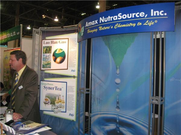 A man in a brown suit stands at the Amax NutraSource, Inc. booth, with posters for Luo Han Guo and SynerTea and a blue backdrop showing water droplets.