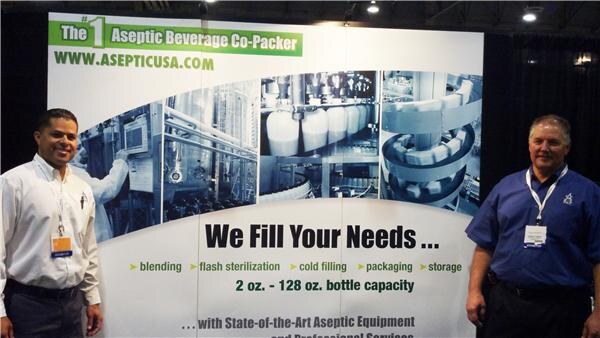 Two men with exhibitor badges stand on either side of an Aseptic USA display reading We Fill Your Needs, with photos of beverage bottling equipment and a list of services.