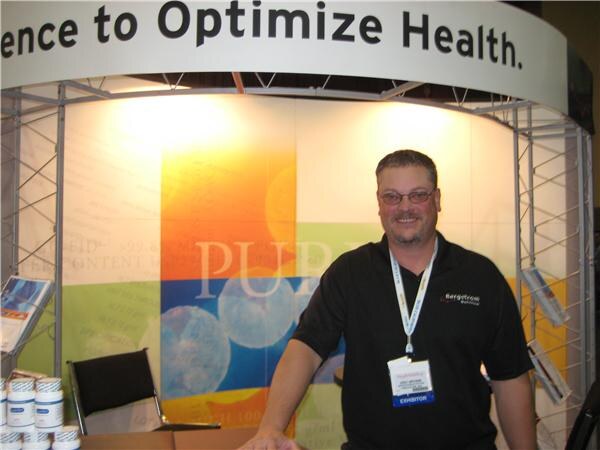 A smiling bearded man with glasses and an exhibitor badge, wearing a black Bergstrom Nutrition shirt, stands in a booth with supplement jars on a table at the lower left.