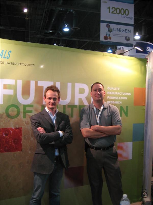 Two men with arms crossed stand in front of a green banner with partial text "Future" and "Quality, Manufacturing, Formulation, Research." Aisle signs for Virgo and Unigen hang overhead.