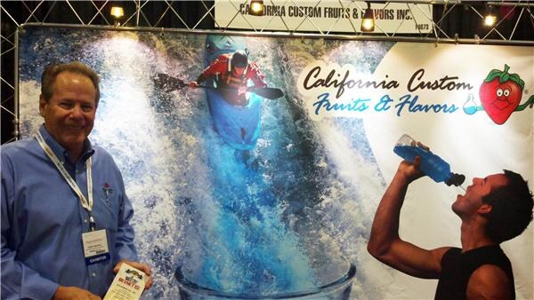 A smiling man in a blue shirt and exhibitor badge holds a brochure in front of a California Custom Fruits & Flavors backdrop showing a kayaker in rapids and a man drinking from a bottle.