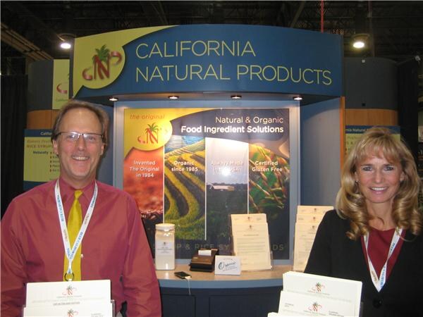 A man in a red shirt and yellow tie and a woman with long blonde hair stand at the California Natural Products booth, which has a sign reading Natural & Organic Food Ingredient Solutions.